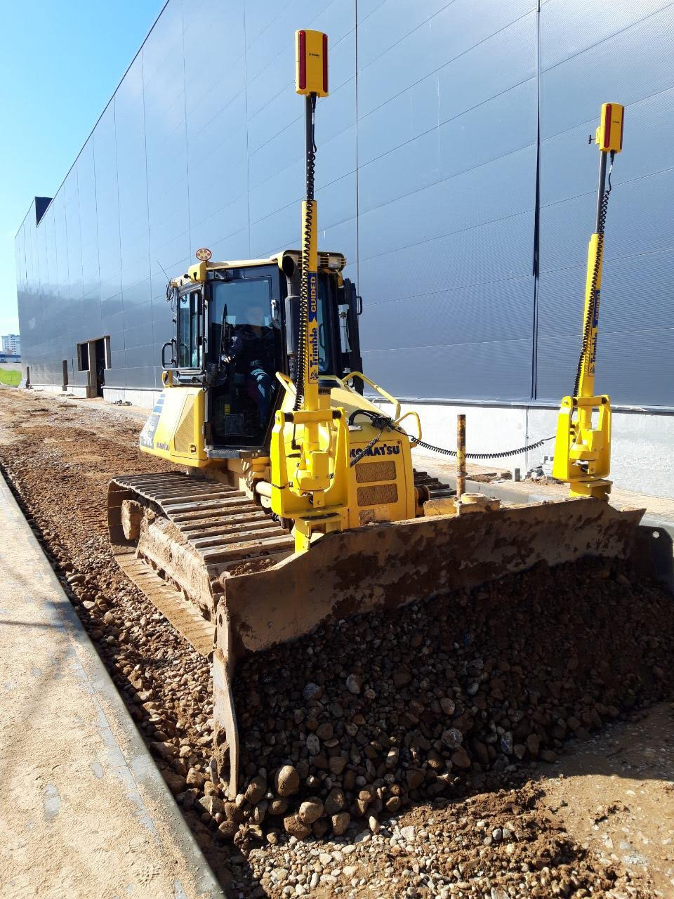 Żółty spychacz Komatsu z systemem Trimble Guided wyrównuje teren przy nowoczesnym budynku przemysłowym, widoczna pryzma ziemi przed lemieszem.