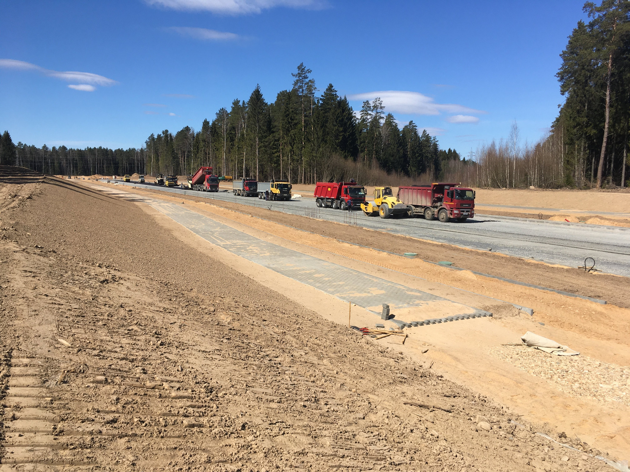 Ciężarówki i walec drogowy pracują na budowie nowej drogi, widoczne przygotowane podłoże, fragmenty kostki brukowej i otaczający las.