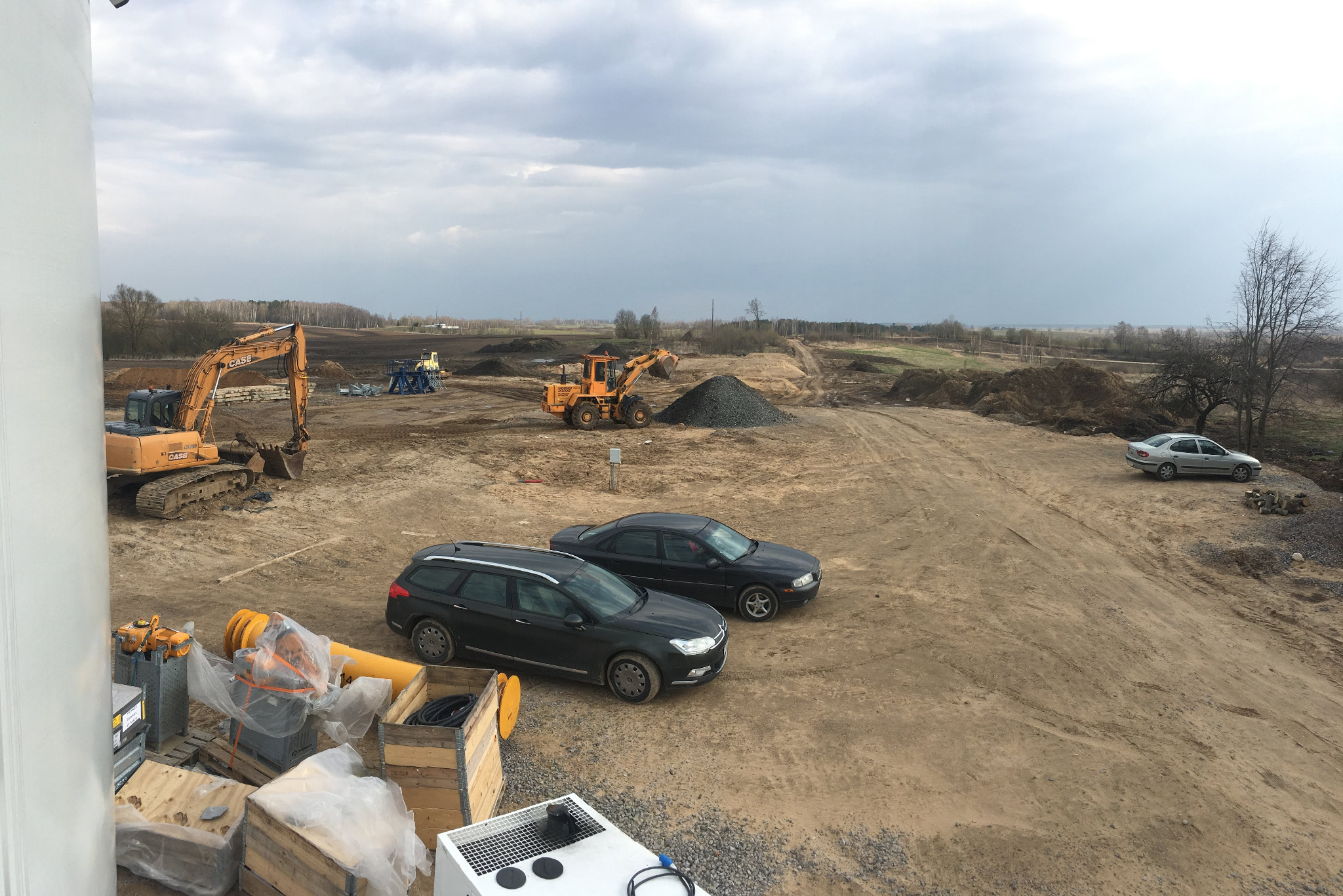 Szeroki plan terenu budowy z dwoma koparkami, stertą żwiru, dwoma zaparkowanymi samochodami osobowymi i elementami konstrukcyjnymi w skrzyniach na pierwszym planie, sfotografowany w pochmurny dzień.