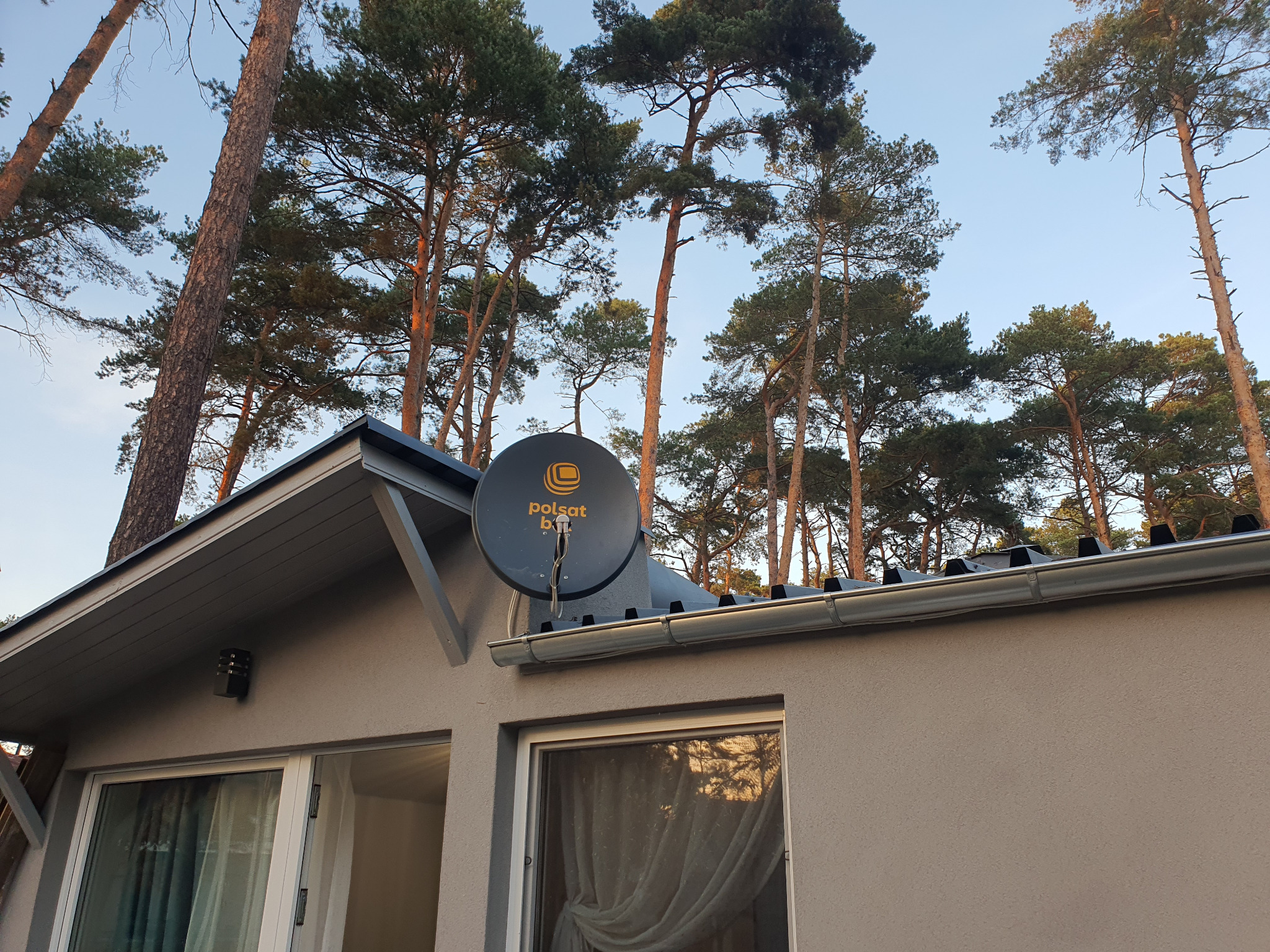 Antena satelitarna Polsat Box zamontowana na ścianie domu z widocznymi drzewami w tle.