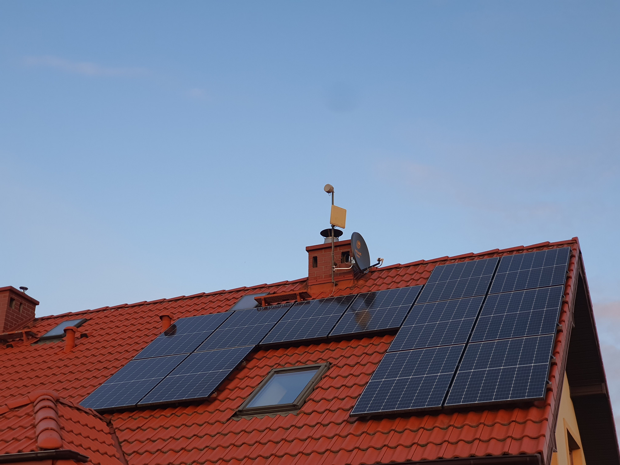 Czerwony dach pokryty dachówką z zamontowanymi panelami fotowoltaicznymi i anteną satelitarną Polsat, widoczne okno dachowe i komin.