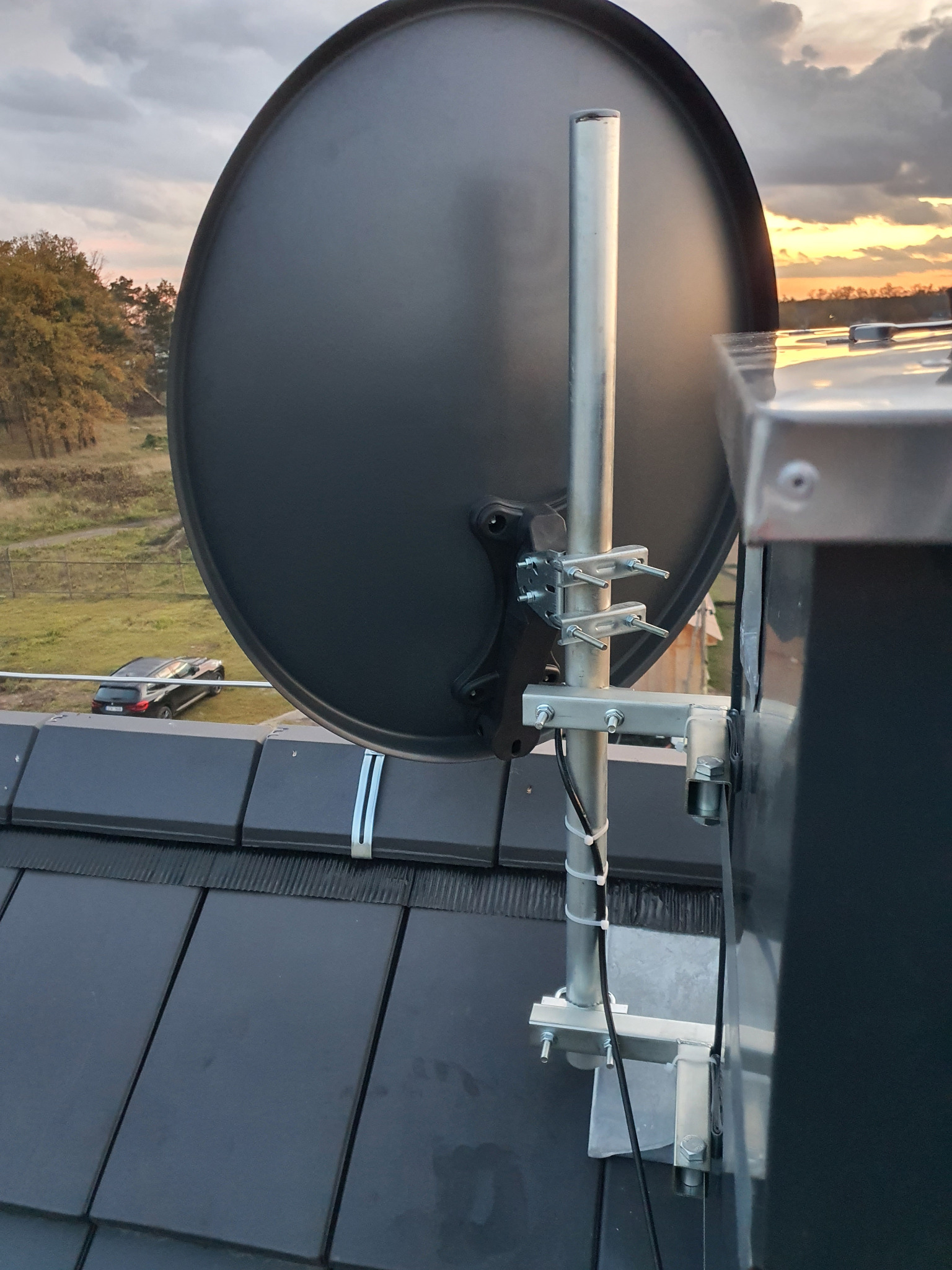 Czarna antena satelitarna zamontowana na dachu z czarnej dachówki, z widocznymi srebrnymi elementami mocującymi i kablem, na tle zachodzącego słońca.