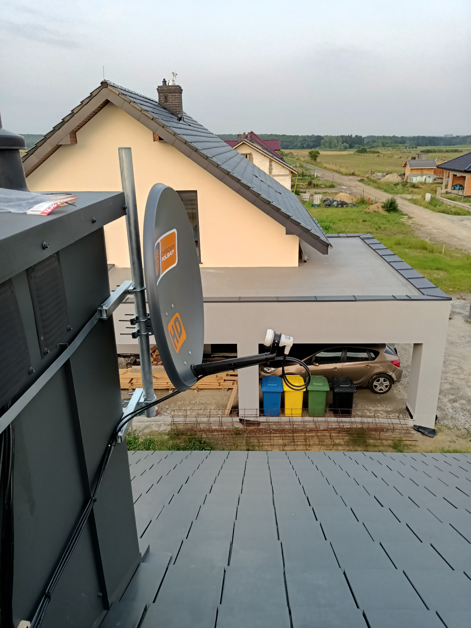 Szara antena satelitarna z logo Cyfrowy Polsat zamontowana na dachu budynku z widokiem na dom jednorodzinny i garaż z samochodem.