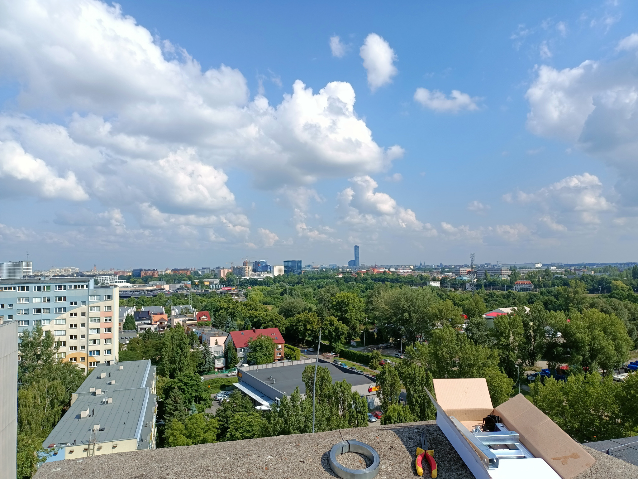 Sprzęt do instalacji na dachu budynku z widokiem na panoramę miasta w tle, widać narzędzia i elementy montażowe w otwartym kartonie.