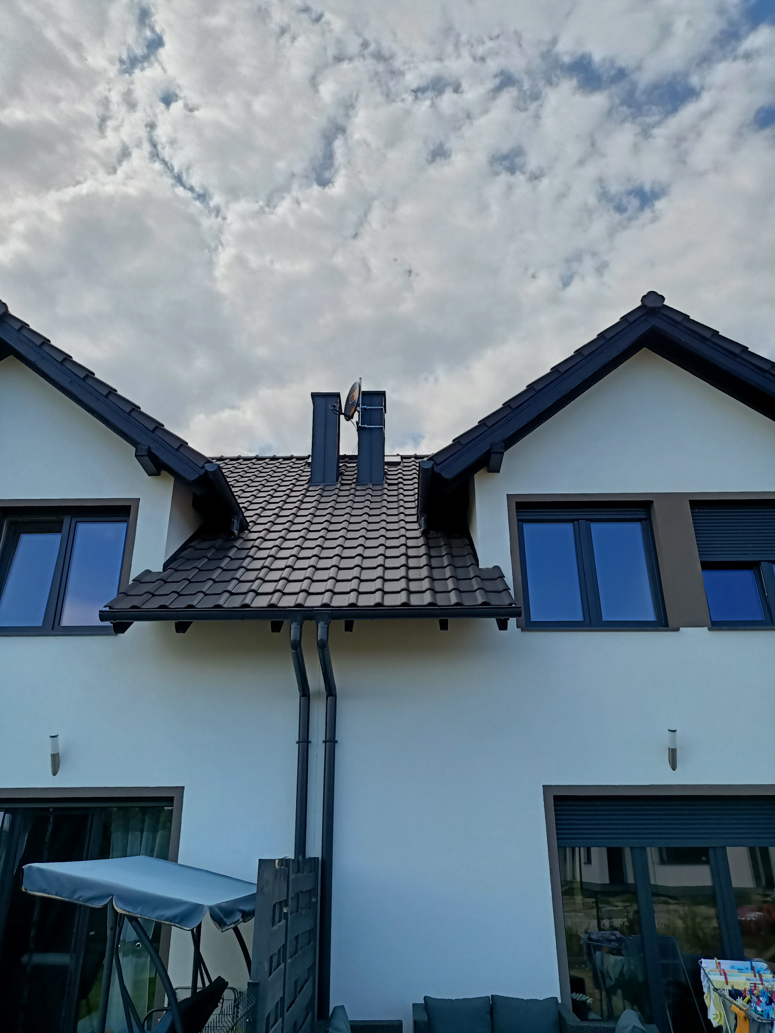 Antena satelitarna zamontowana na kominie nowoczesnego domu jednorodzinnego z ciemną dachówką i orynnowaniem, widok z dołu na tle pochmurnego nieba.