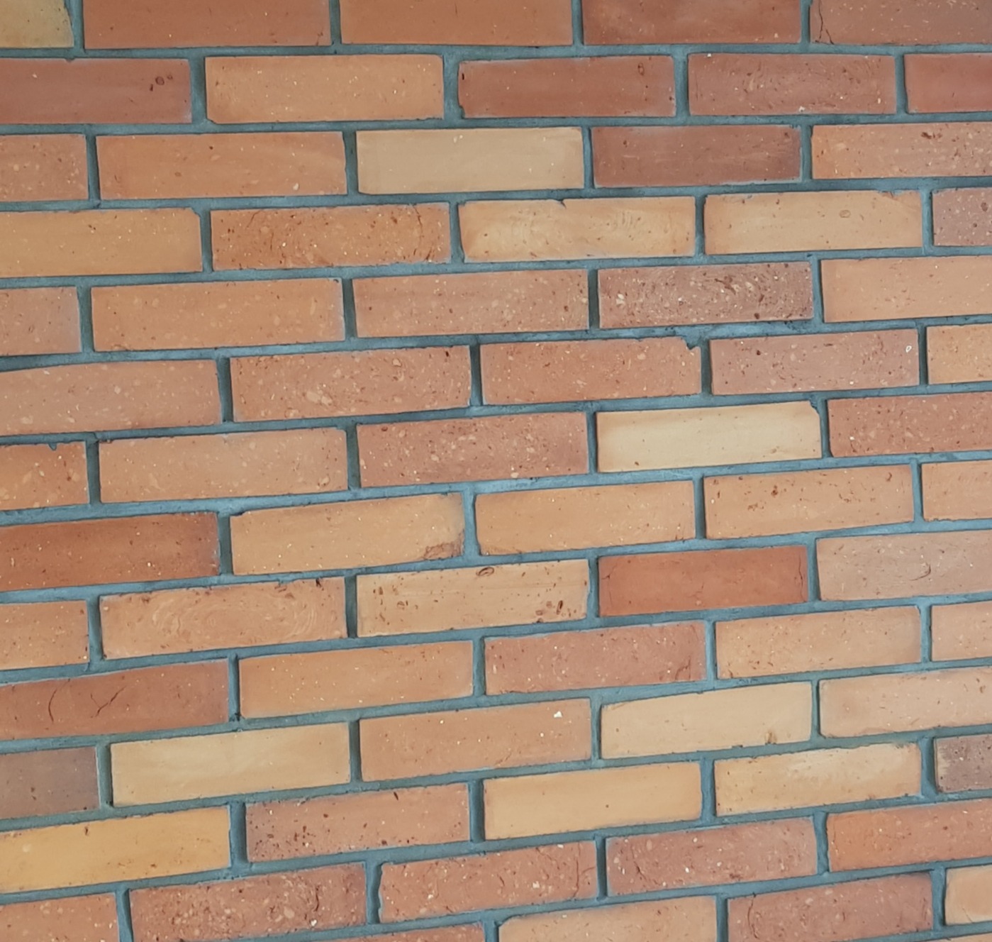 Ściana wyłożona cegłami klinkierowymi w odcieniach czerwieni i pomarańczy, z szarą fugą, ułożona pod kątem.