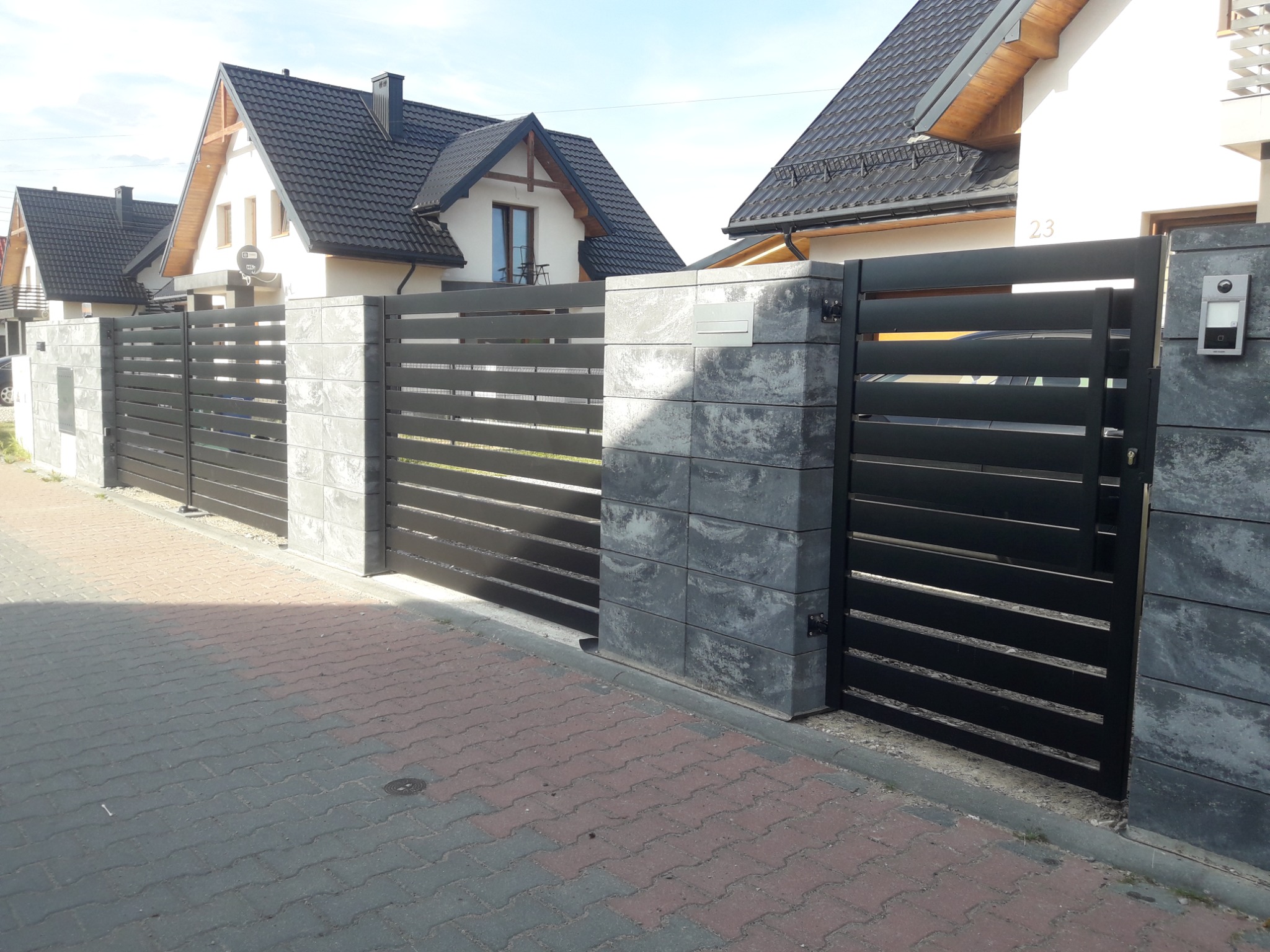 Nowoczesne ogrodzenie posesji z poziomych, czarnych profili metalowych, osadzone na słupkach z szarej, betonowej kostki, na tle domów jednorodzinnych.