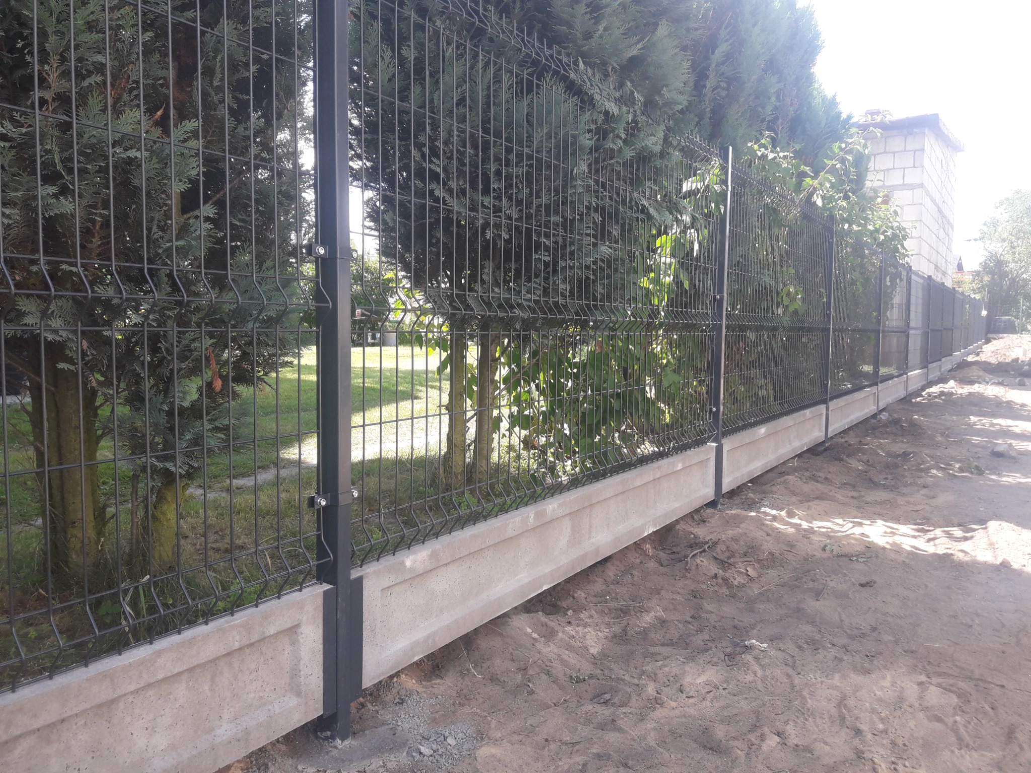 Długa, metalowa siatka ogrodzeniowa panelowa z podmurówką betonową, otoczona zielenią i drzewami.