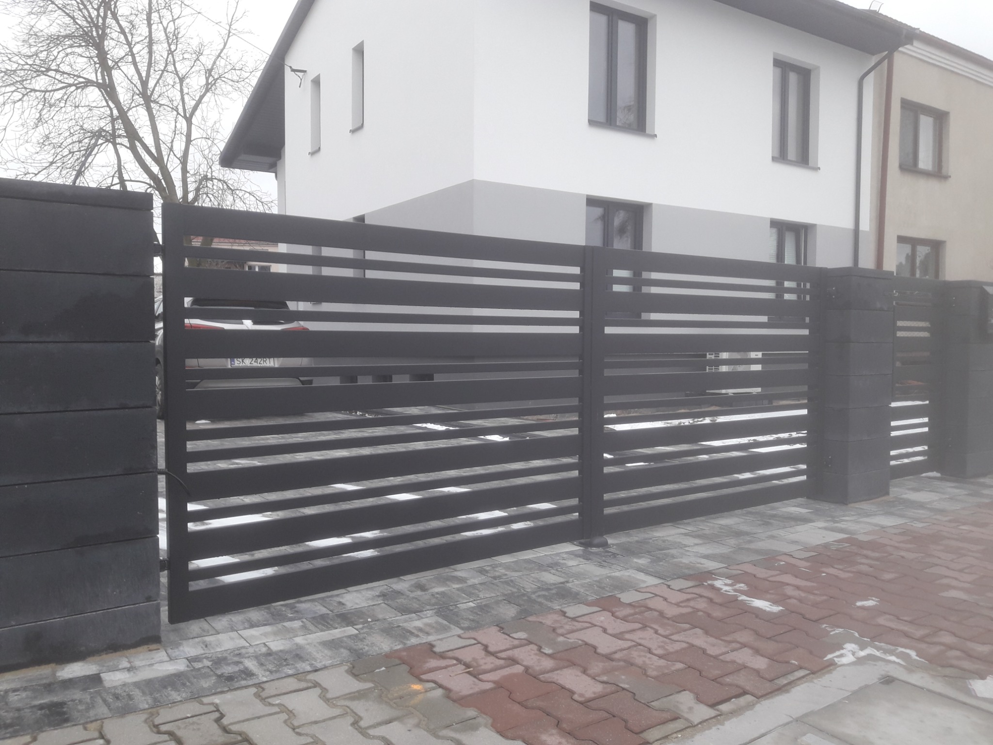 Nowoczesna, przesuwna brama wjazdowa z poziomych, czarnych profili, osadzona na betonowych słupkach, na tle dwukondygnacyjnego domu z białymi ścianami i kostki brukowej.