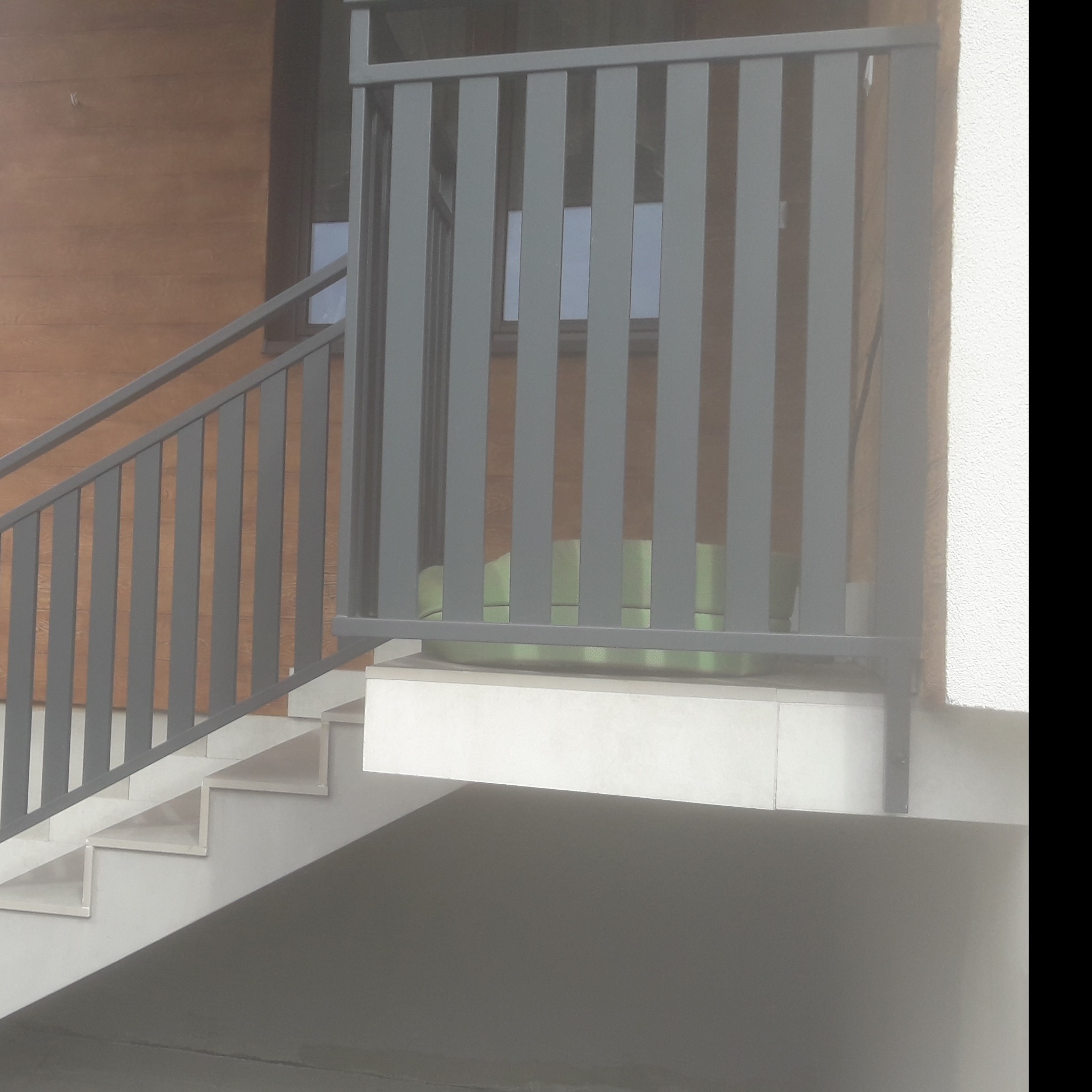 Nowoczesna balustrada schodowa z szarych, pionowych prętów, z widocznymi schodami i fragmentem elewacji budynku z drewnianą okładziną.