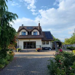 Dom jednorodzinny z brązowym dachem i panelami fotowoltaicznymi, widok z brukowanego podjazdu, bujna roślinność w ogrodzie, drabina oparta o ścianę budynku.