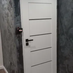 Białe drzwi wewnętrzne z poziomymi frezowaniami i czarną klamką, zamontowane w ścianie z dekoracyjnym, ciemnym tynkiem strukturalnym, widoczne elementy instalacji elektrycznej po lewej stronie.