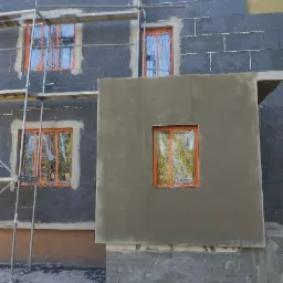 Ocieplanie elewacji budynku z widocznymi rusztowaniami, oknami zabezpieczonymi folią i fragmentem ściany po nałożeniu szarego tynku.