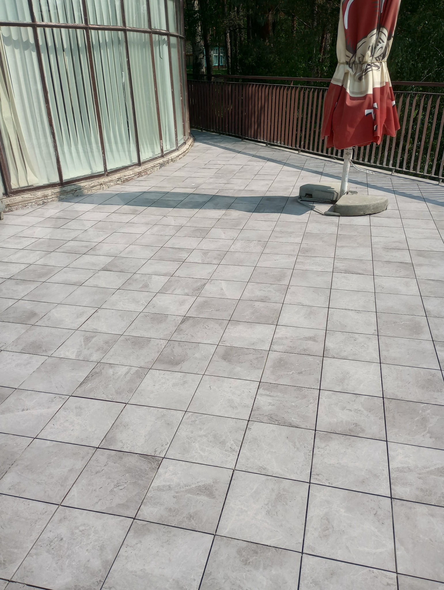 Ułożona szara, kwadratowa terakota na tarasie z widokiem na przeszkloną werandę i częściowo złożony parasol ogrodowy.