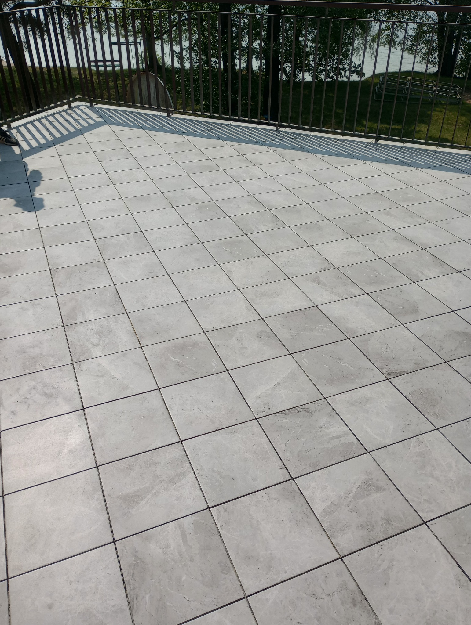 Wyłożony jasnoszarą marmurkową płytką taras z metalową barierką i widokiem na zieleń i wodę w tle.