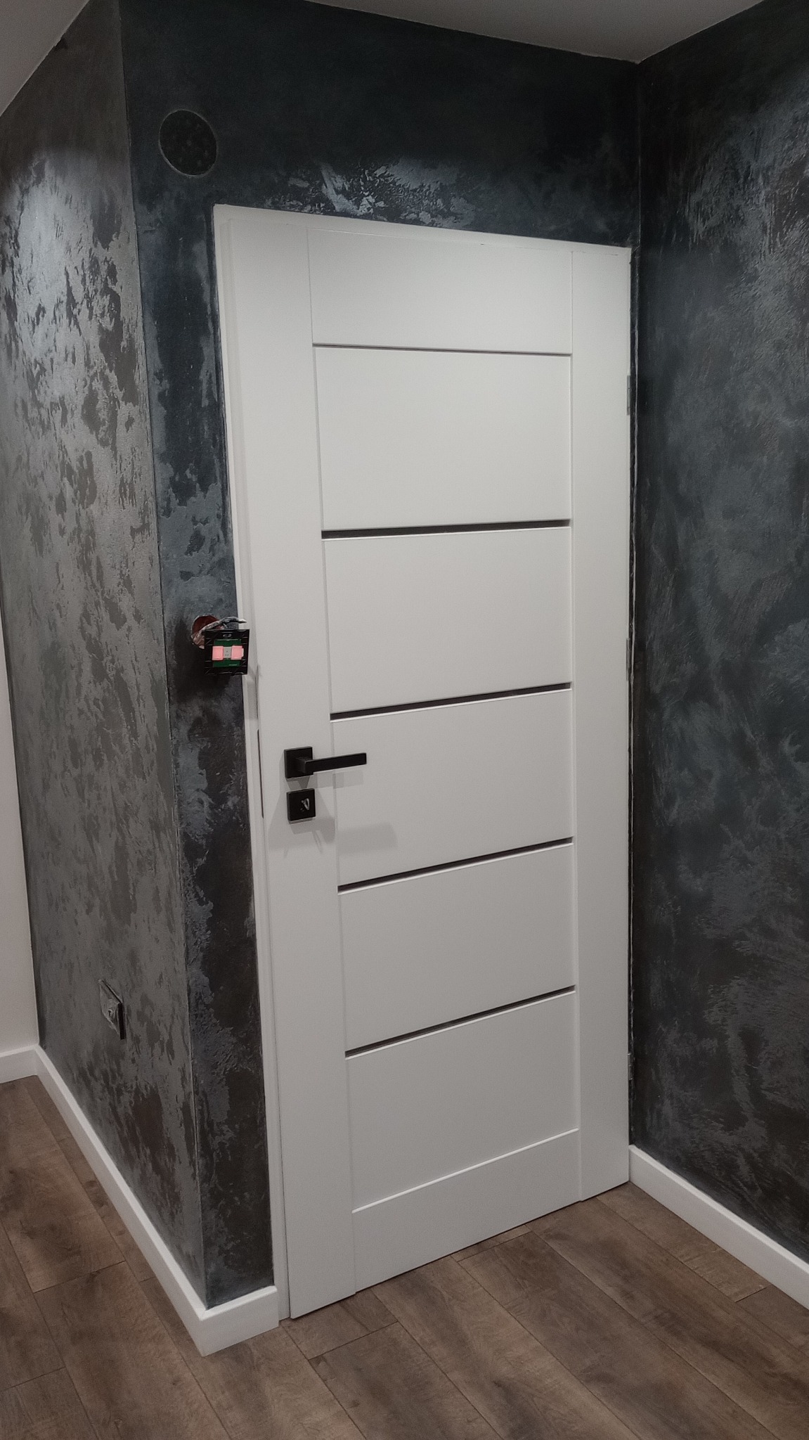 Białe drzwi wewnętrzne z poziomymi frezowaniami i czarną klamką, zamontowane w ścianie z dekoracyjnym, ciemnym tynkiem strukturalnym, widoczne elementy instalacji elektrycznej po lewej stronie.