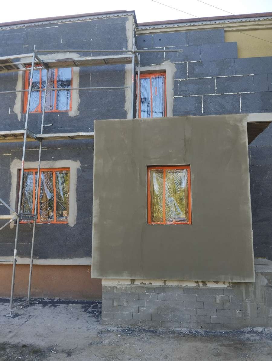 Ocieplanie elewacji budynku z widocznymi rusztowaniami, oknami zabezpieczonymi folią i fragmentem ściany po nałożeniu szarego tynku.