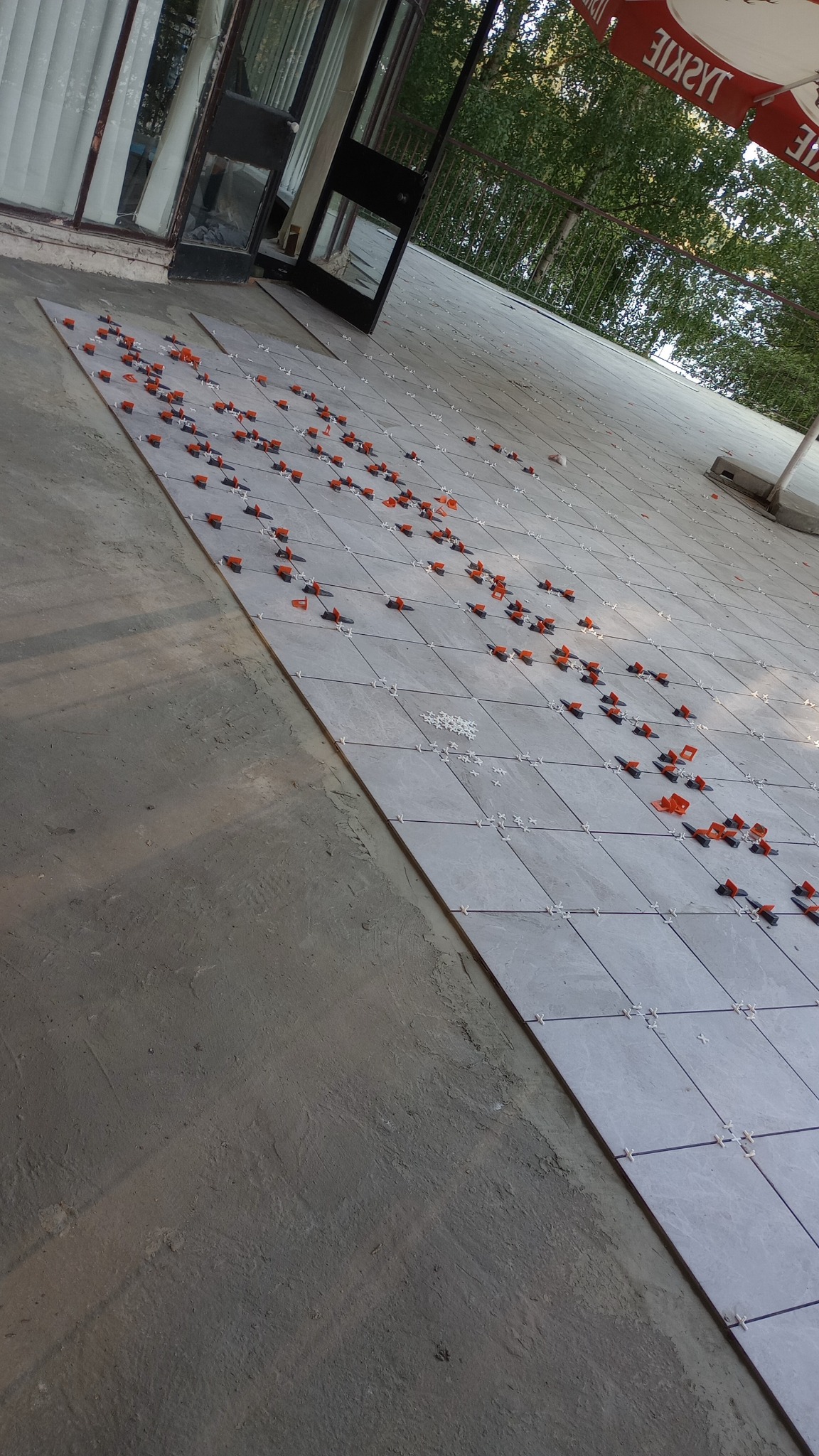 Układanie szarych płytek na zewnątrz budynku, widoczne pomarańczowe klipsy poziomujące i białe krzyżyki dystansowe. Prace w toku, widoczny fragment surowego podłoża i parasol reklamowy w tle.