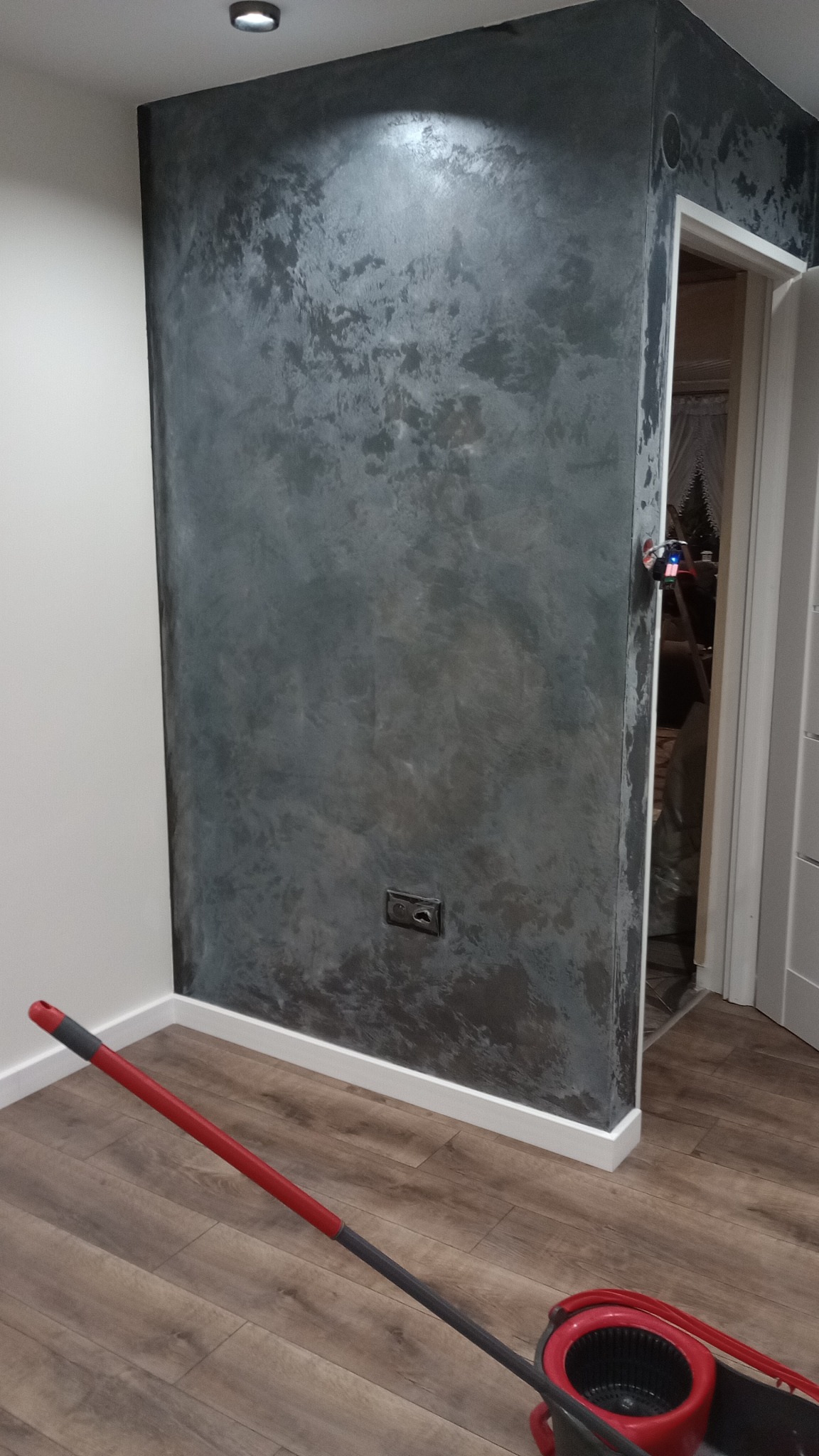 Ściana z dekoracyjnym efektem betonu w odcieniach szarości, widoczny kontakt elektryczny, mop z czerwonym wiadrem na drewnianej podłodze, fragment białej listwy przypodłogowej i ościeżnicy drzwiowej.