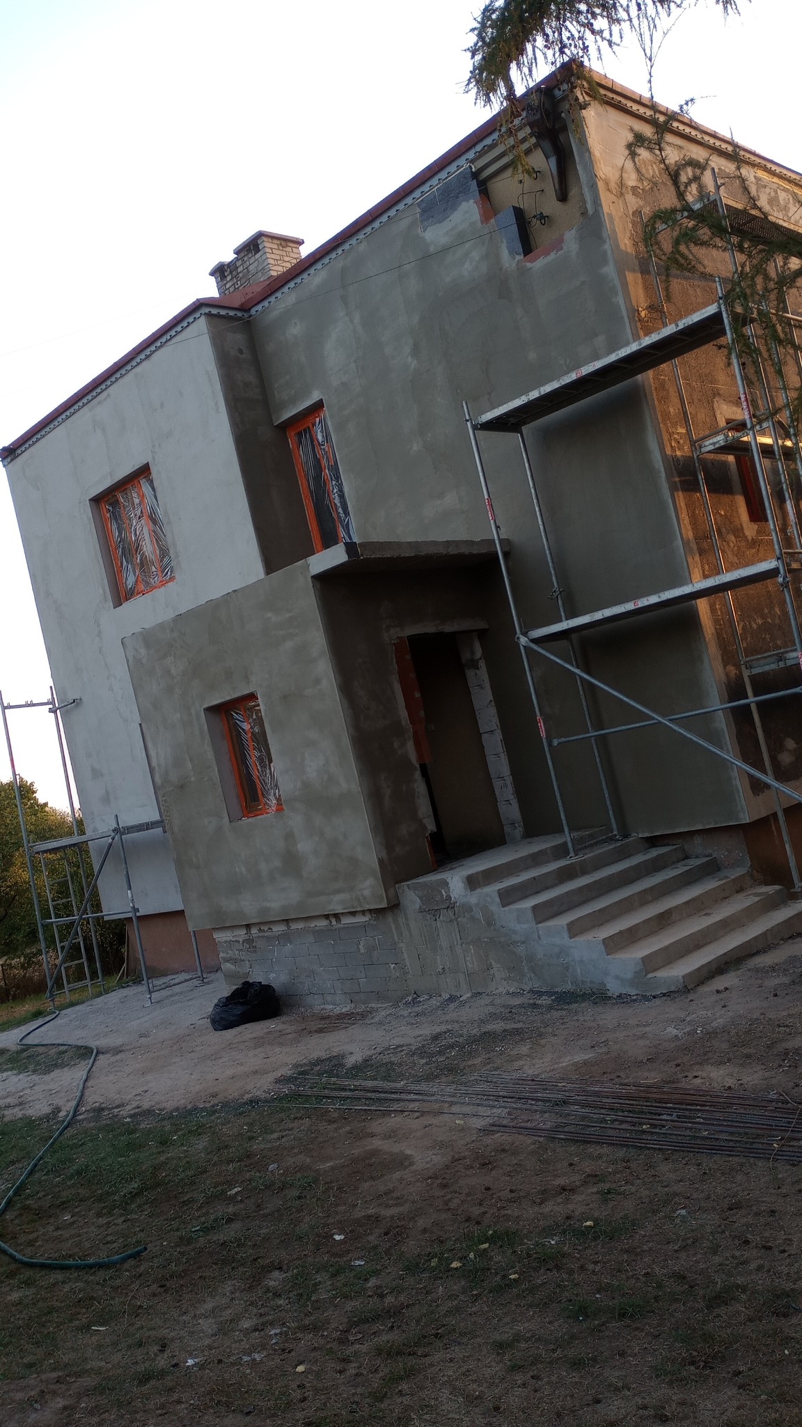 Dwupiętrowy dom w trakcie ocieplania elewacji, z oknami zabezpieczonymi folią, otoczony rusztowaniami i materiałami budowlanymi.