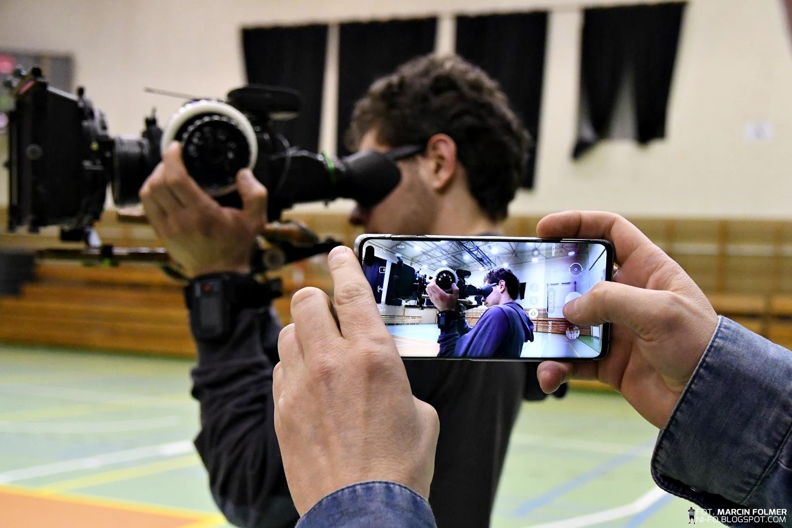 Mężczyzna filmujący kamerą na statywie, uchwycony na ekranie smartfona trzymanego w dłoniach innej osoby, w tle hala sportowa.