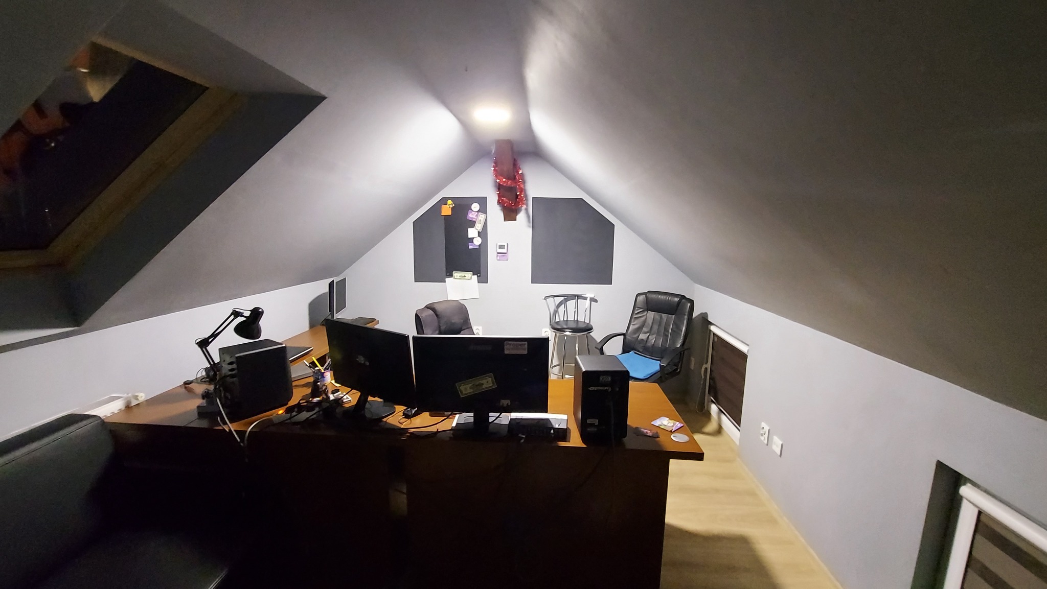 Biuro na poddaszu z trzema monitorami, komputerem stacjonarnym, lampką biurkową i dwoma krzesłami, oświetlone centralnym punktem świetlnym i oknem dachowym.