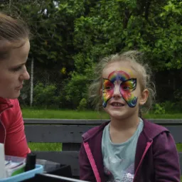 Dziewczynka z pomalowaną twarzą w motywy tęczowego motyla, siedząca na ławce obok kobiety z różową kokardą we włosach, która patrzy na nią z uwagą.