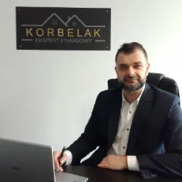 Mężczyzna w garniturze siedzi przy biurku z laptopem, trzyma długopis w dłoni, za nim widoczna tabliczka z napisem KORBELAK Ekspert Finansowy i logotypem.
