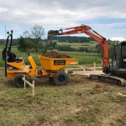 Pomarańczowy minikoparka nabiera ziemię do żółtej wywrotki na tle zielonego pola i pagórków pod pochmurnym niebem, widoczne drewniane szalunki.