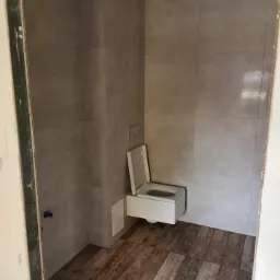 Nowoczesna toaleta podwieszana z otwartą klapą, szare płytki ścienne i drewnopodobne płytki podłogowe, widok na wykończone wnętrze łazienki z listwą sufitową.