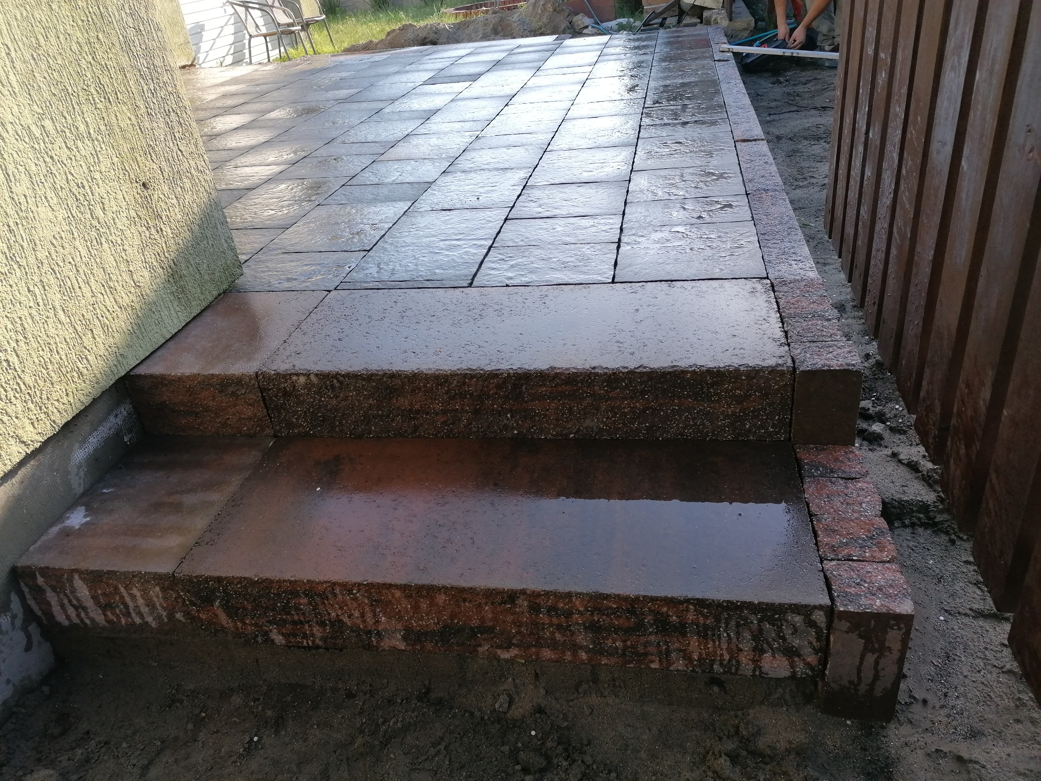 Ułożone schody wejściowe z kostki granitowej w kolorze czerwono-szarym, mokre od deszczu, z widocznym fragmentem brukowanej ścieżki i drewnianego ogrodzenia w tle.
