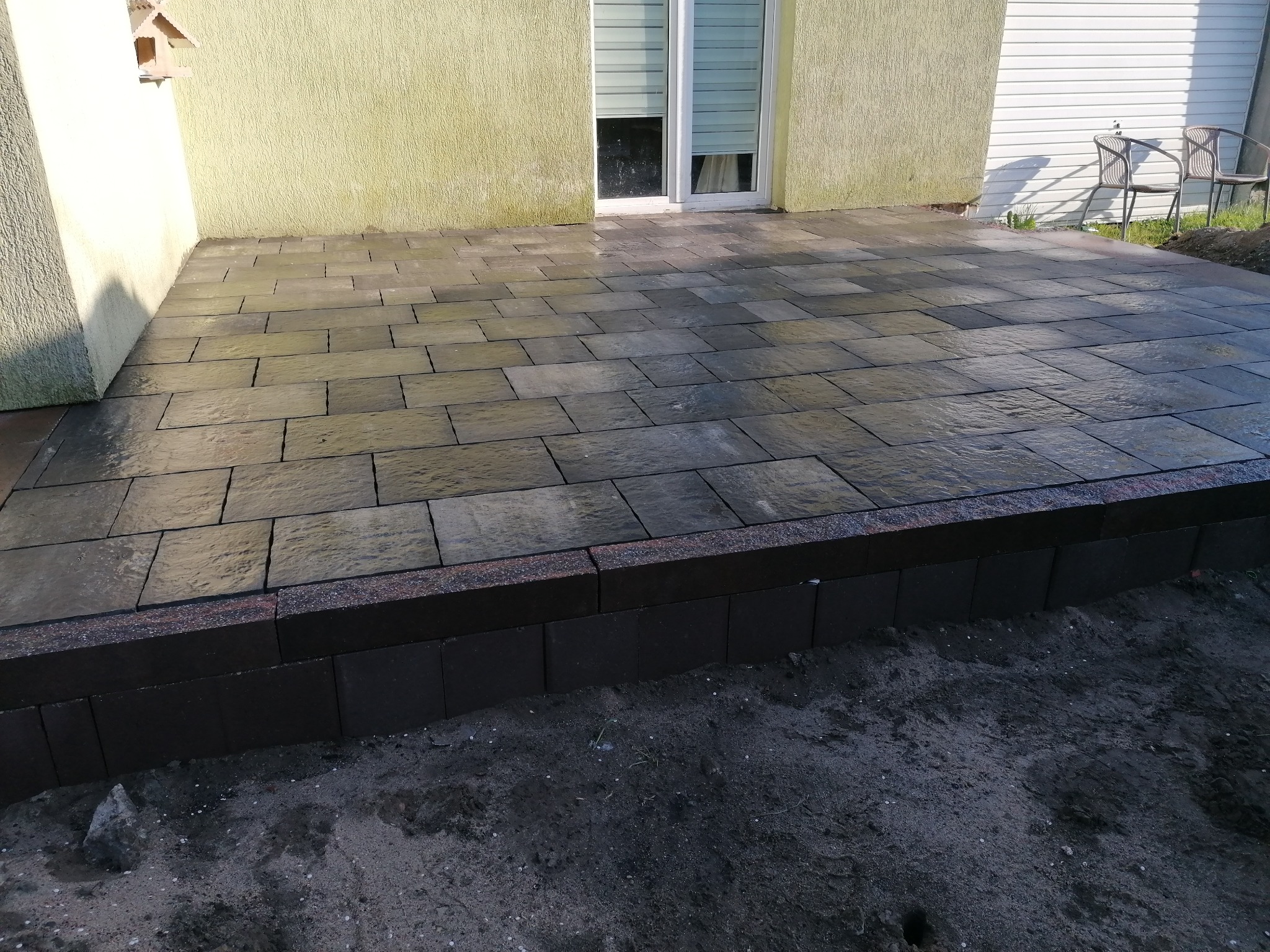 Świeżo ułożona, mokra nawierzchnia z ciemnoszarej kostki brukowej, obrzeże z ciemnej kostki granitowej, widok na ścianę budynku z oknem i dwa krzesła ogrodowe w tle.