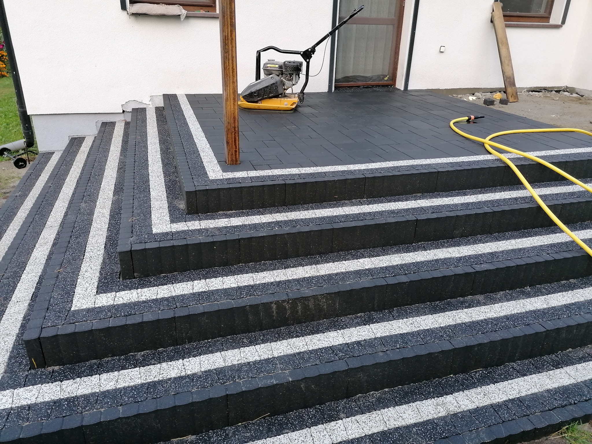 Schody wejściowe do domu wyłożone czarną kostką brukową z białymi akcentami, widoczna zagęszczarka i żółty wąż ogrodowy.