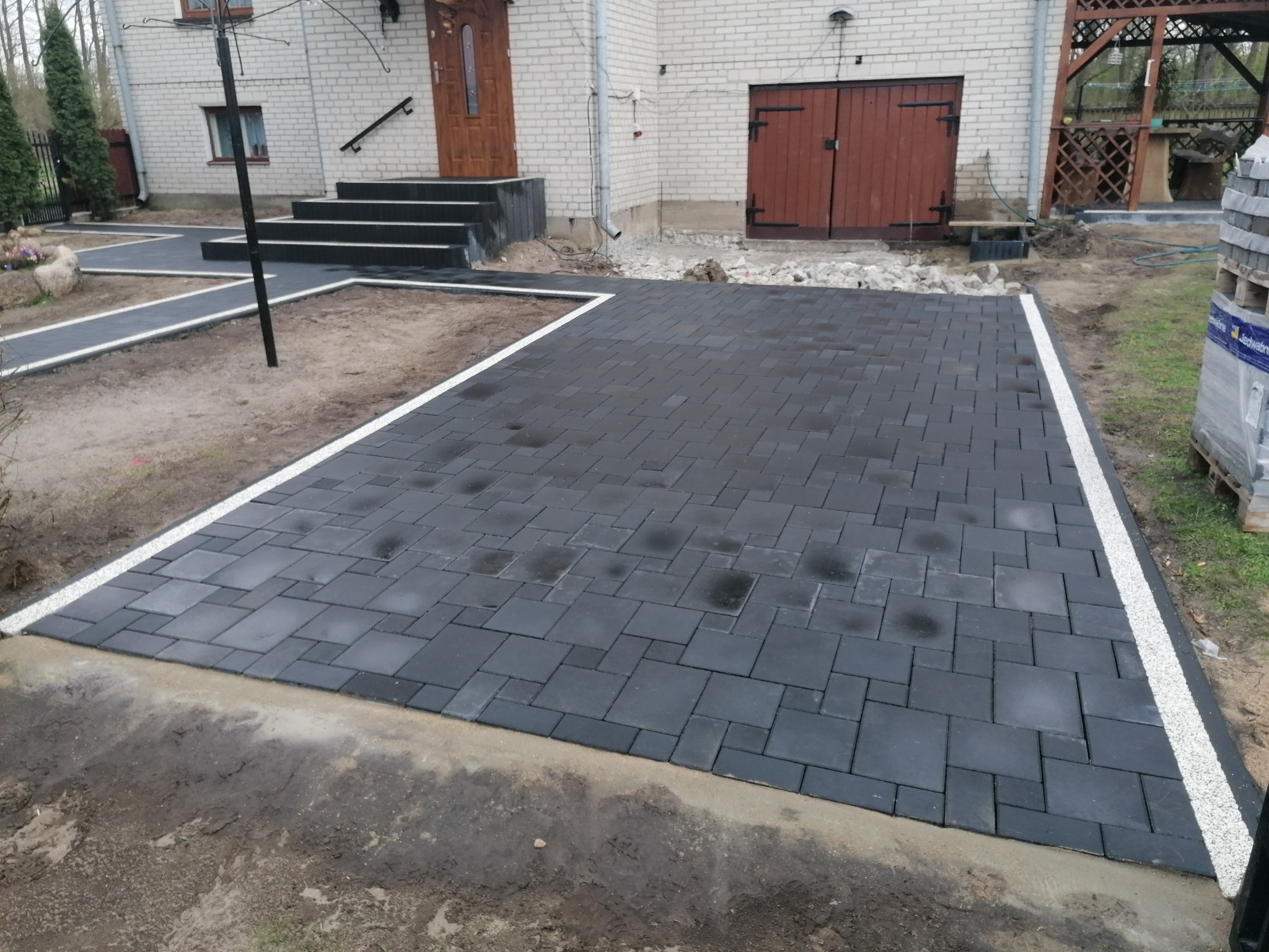 Ukończony podjazd z ciemnej kostki brukowej ułożonej w nieregularny wzór, obrzeża z białego grysu, tło stanowi budynek z cegły i elementami drewnianymi.