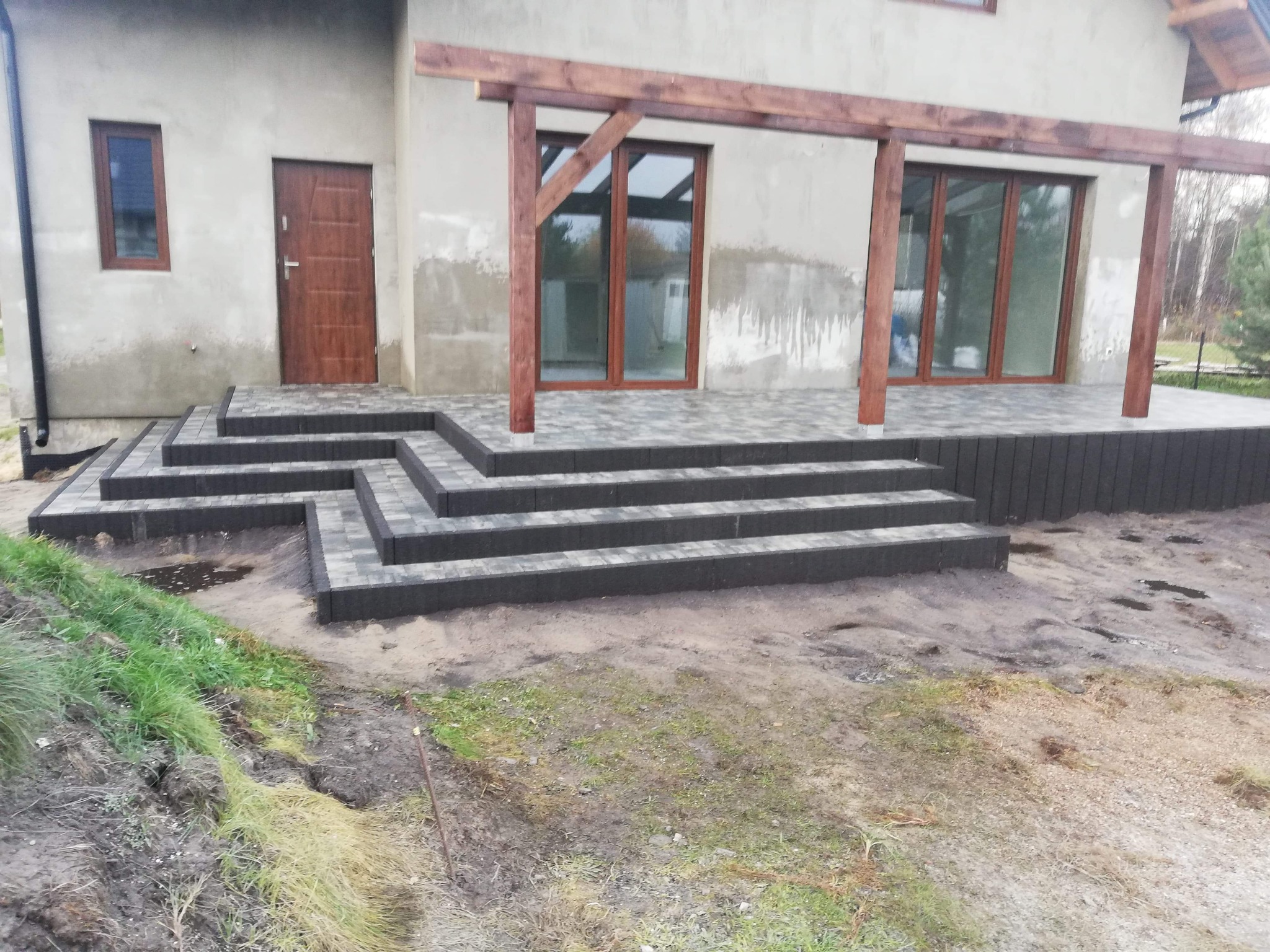 Schody wejściowe z szarej kostki brukowej prowadzące do domu z drewnianą pergolą, widoczne ślady prac ziemnych.