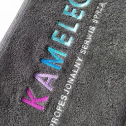 Szary ręcznik frotte z wyhaftowanym kolorowym napisem 'KAMELEON' i białym napisem 'PROFESJONALNY SERWIS SPRZĄTAJĄCY'.