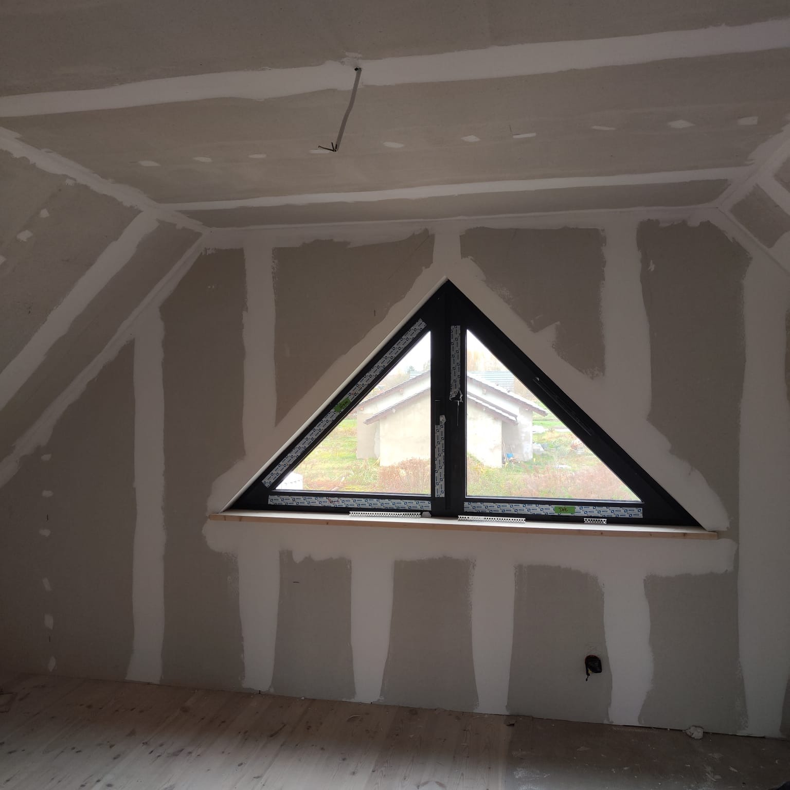 Poddasze z trójkątnym oknem po szpachlowaniu, widoczne łączenia płyt gipsowych. Surowe drewno podłogi i kabel zwisający z sufitu dodają surowości wnętrzu.