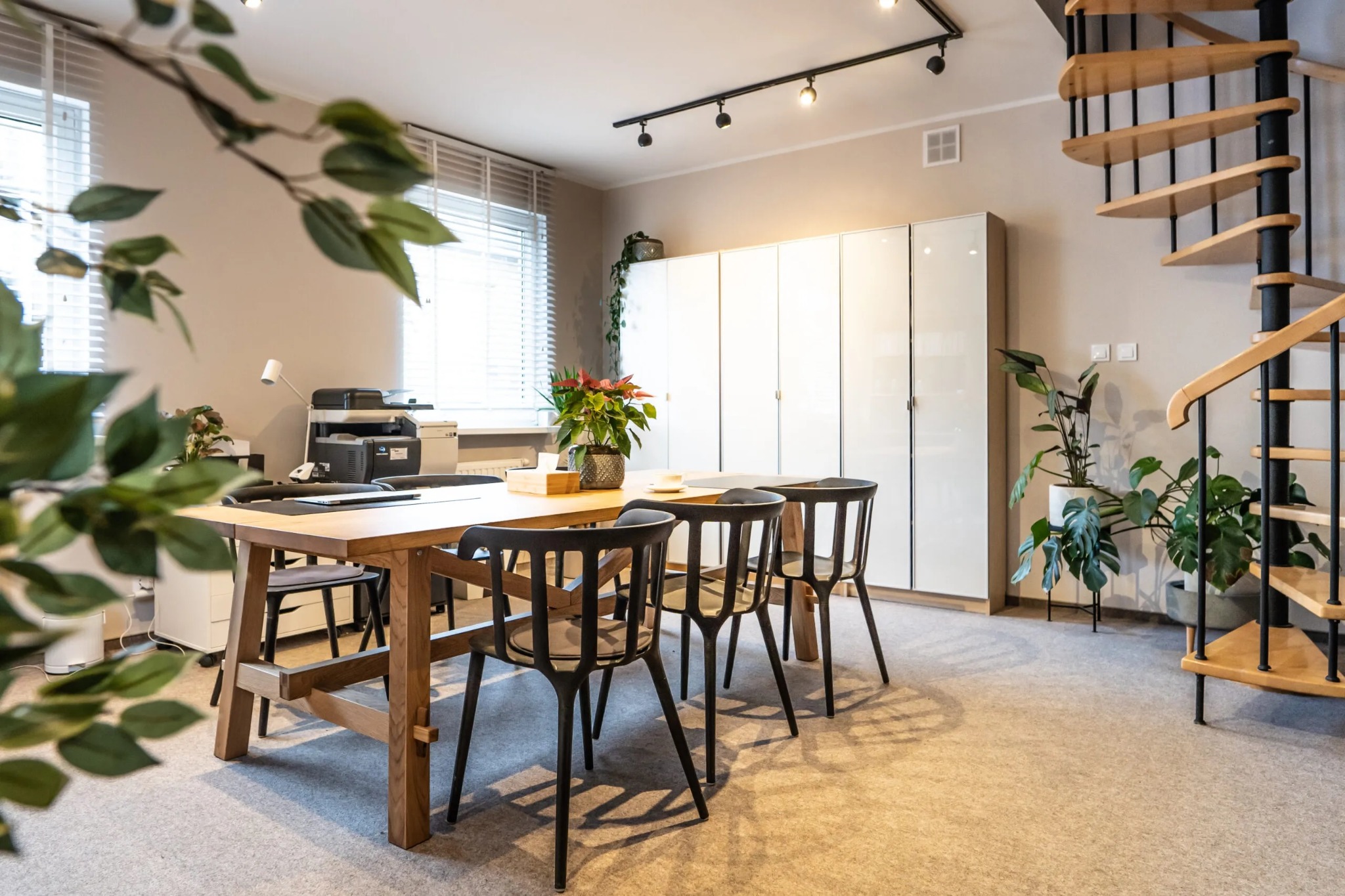 Jasne biuro z drewnianym stołem, czarnymi krzesłami, spiralnymi schodami i roślinami doniczkowymi, widok zza zielonych gałęzi