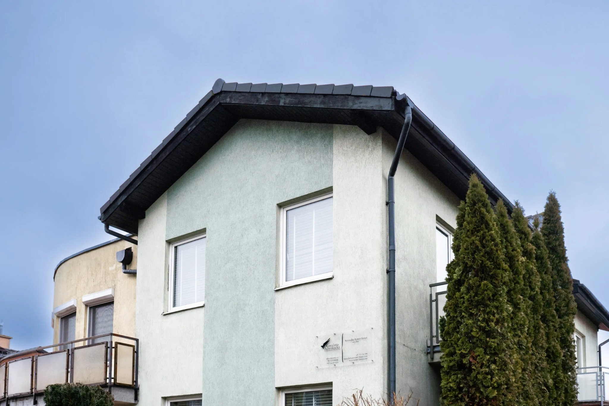 Dwupiętrowy budynek z szarą elewacją, ciemnym dachem i rynnami, z tabliczką informacyjną przy wejściu i wysokimi tujami po prawej stronie, widziany z dołu pod pochmurnym niebem.