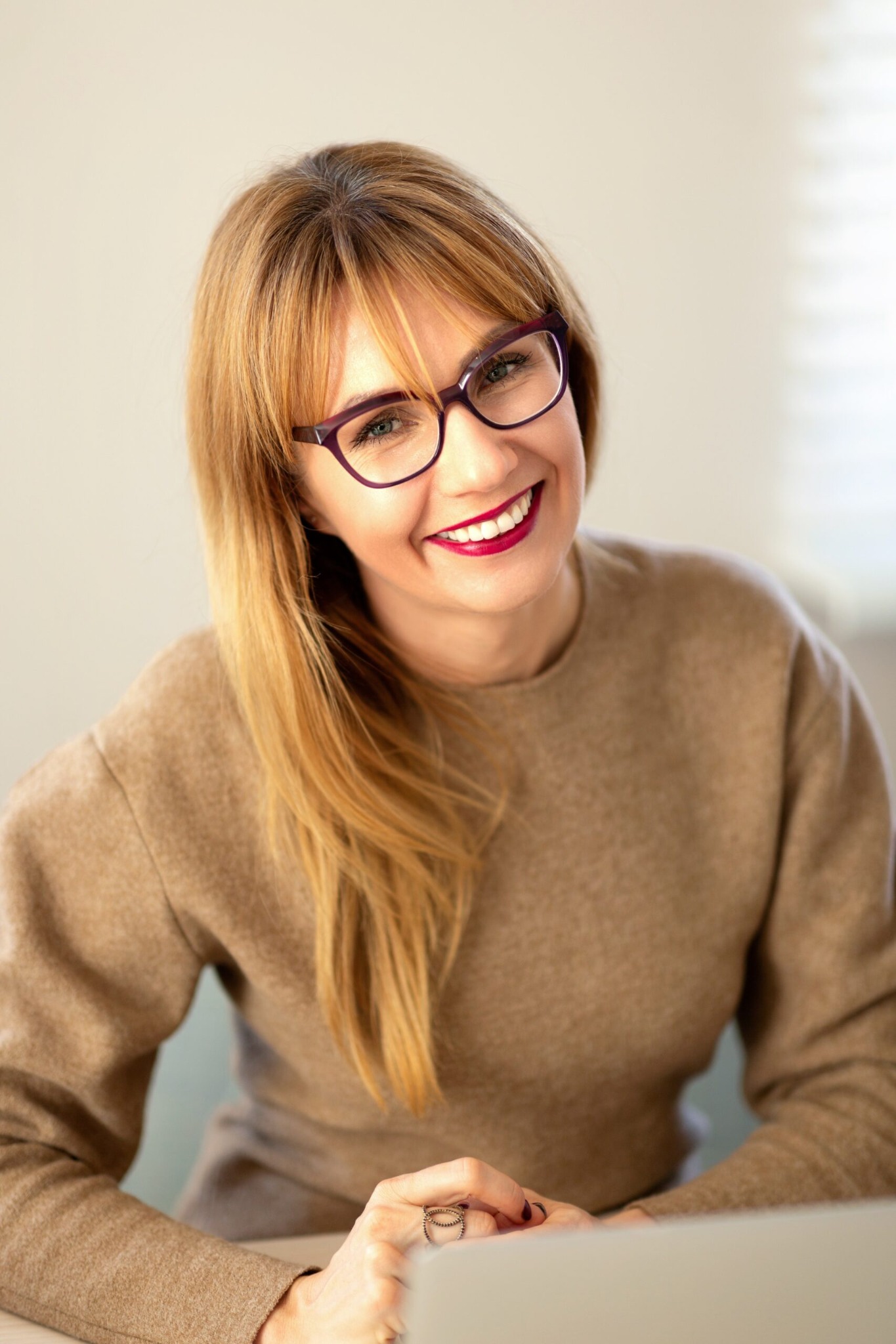 Uśmiechnięta kobieta z blond włosami i okularami w bordowych oprawkach, ubrana w brązowy sweter, siedzi przed laptopem. Na palcu prawej ręki ma pierścionek z ozdobnymi kamieniami.