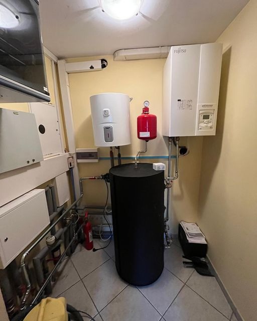 Instalacja grzewcza z czarnym zbiornikiem akumulacyjnym, białym bojlerem i czerwonym naczyniem wzbiorczym w kotłowni w Poznaniu, widoczna część rur i urządzeń sterujących.