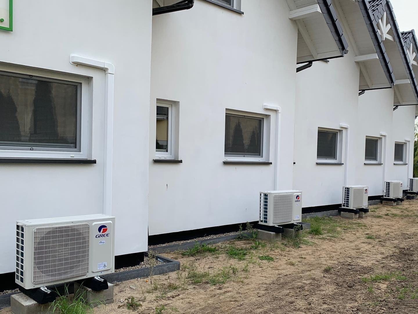 Rząd identycznych, białych jednostek zewnętrznych klimatyzacji marki Gree, zamontowanych na betonowych podstawach przy ścianach szeregowca z białymi elewacjami i czarnymi dachami.