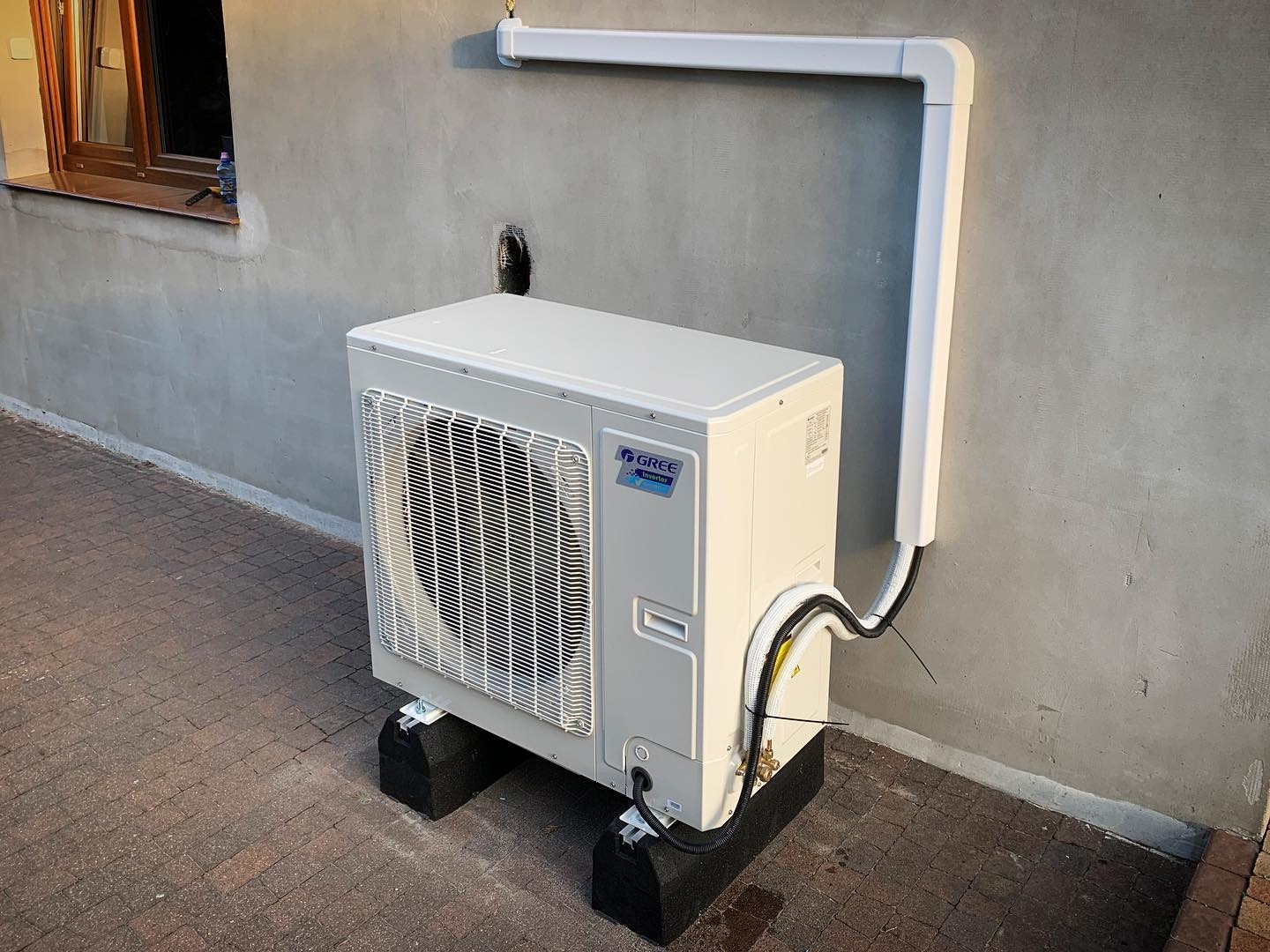 Zewnętrzna jednostka klimatyzacji marki Gree zamontowana na czarnych podstawach antywibracyjnych przy szarej ścianie budynku z widocznymi przewodami i rurami odprowadzającymi.
