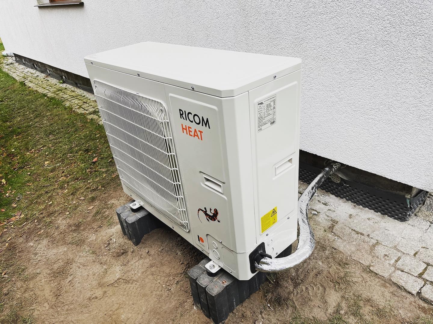 Biała pompa ciepła Ricom Heat zamontowana na betonowych blokach przy ścianie budynku z szorstkim tynkiem. Rura odprowadzająca owinięta srebrną folią izolacyjną. Na obudowie naklejka z jaszczurką.