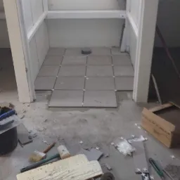 Montaż szarych płytek podłogowych w małym pomieszczeniu, widoczne narzędzia: szlifierka kątowa, miarka, poziomica, krzyżyki dystansowe i fragmenty płytek.