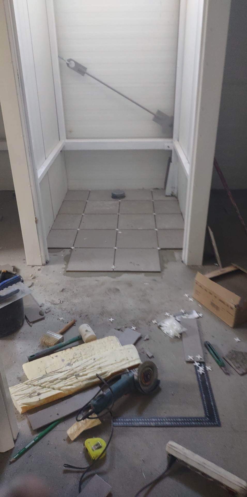 Montaż szarych płytek podłogowych w małym pomieszczeniu, widoczne narzędzia: szlifierka kątowa, miarka, poziomica, krzyżyki dystansowe i fragmenty płytek.