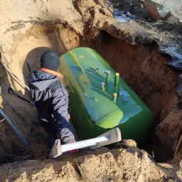 Zielony zbiornik na gaz zakopywany w wykopie, widoczny pracownik w kombinezonie i drabina, ziemia piaszczysta, w tle częściowo widoczny sprzęt budowlany i ogrodzenie.