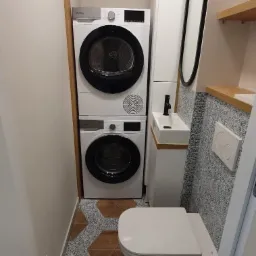 Mała łazienka z pralką i suszarką w zabudowie, umywalką nablatową, wiszącą toaletą i wzorzystą podłogą w kształcie heksagonów z efektem drewna i ornamentów.
