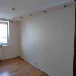 Pomalowane na jasno ściany pokoju z drewnianą podłogą, widoczne lampy sufitowe i okno z parapetem.