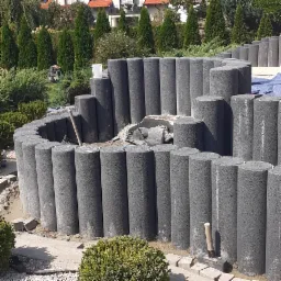 Budowa kaskadowego murku oporowego z betonowych palisad w ogrodzie, widoczne narzędzia i elementy konstrukcyjne.